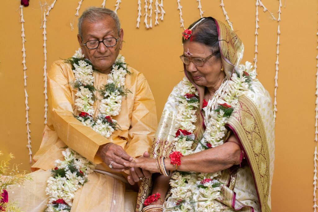 An elderly couple celebrates a traditional ceremony with floral garlands indoors.